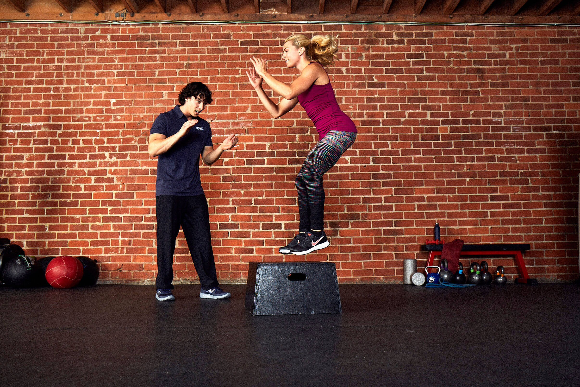 Male NASM trainer assisting female client on box jumps