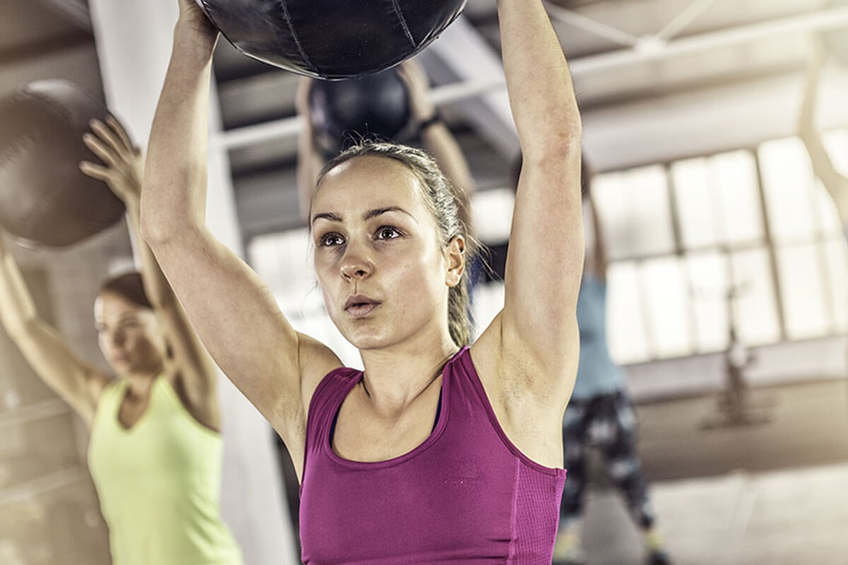Woman holding weighted ball over head in group fitness class
